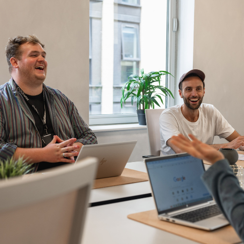 Three people sitting around a table in an office, two of them are laughing. Laptops are open, and a plant is on the window sill in the background.