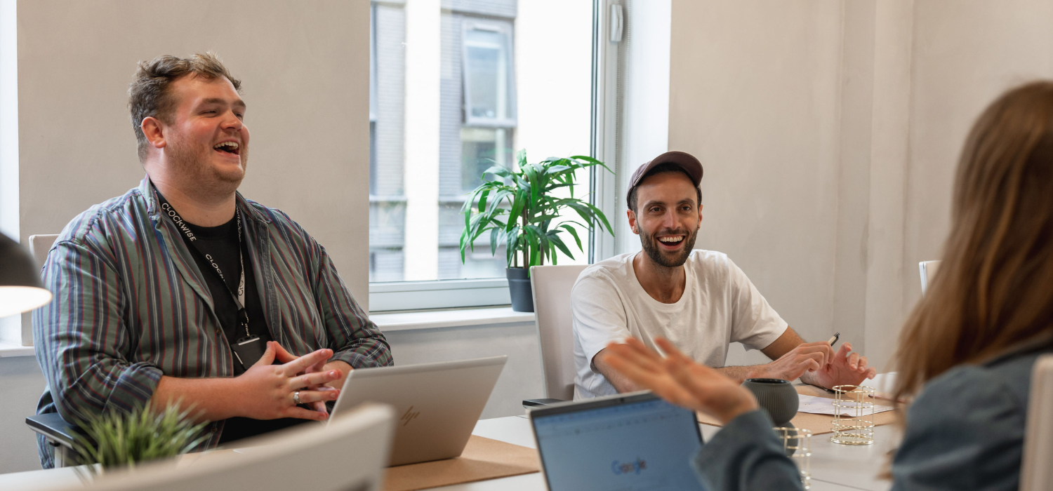 Three people sitting around a table in an office, two of them are laughing. Laptops are open, and a plant is on the window sill in the background.