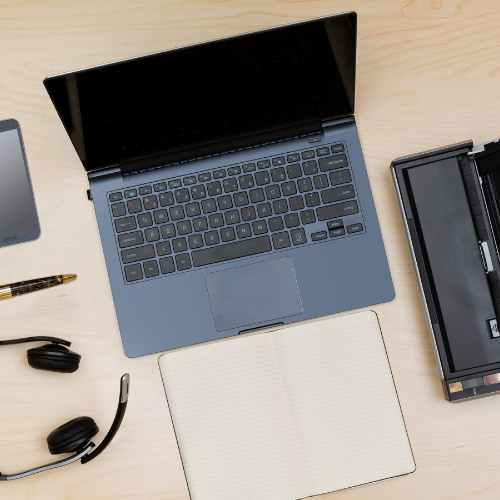 A laptop on a wooden desk with a blank notebook, a headset, a pen, a smartphone, and a partially visible printer. The items are neatly arranged, creating an organized workspace.