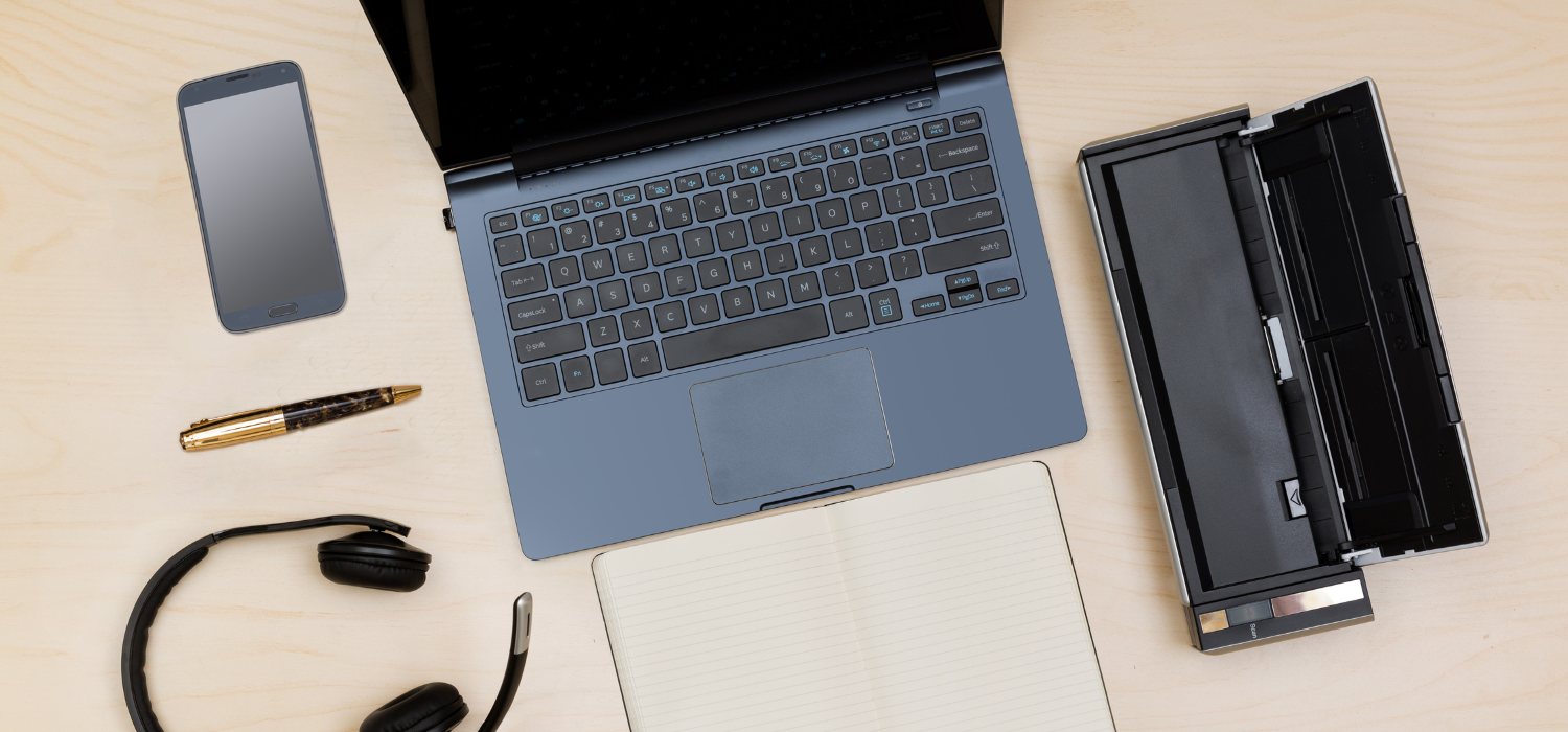 A flat lay of a workspace featuring a laptop, a smartphone, an open notebook, a pen, headphones, and a portable scanner on a light wood surface.