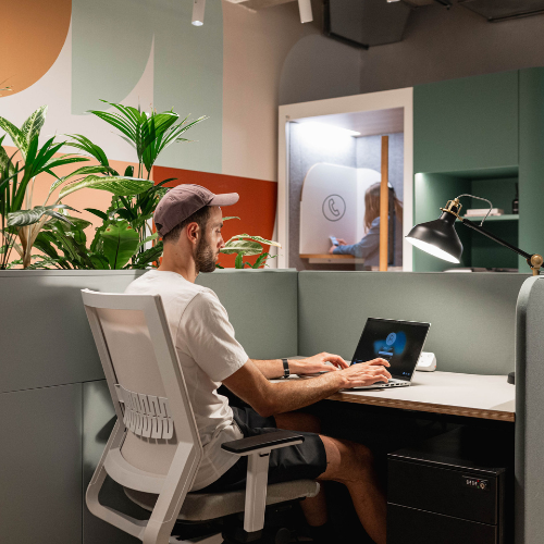 A man in a white shirt and pink cap works on a laptop at a modern desk with plants. A woman is in a small booth behind him, possibly on a call. The space is decorated with pastel colors and has a contemporary design.