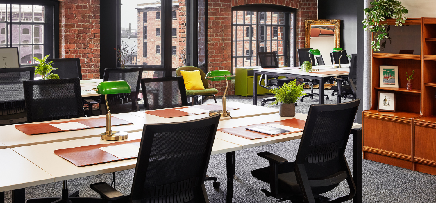 A modern office space with exposed brick walls and large windows. Desks are neatly arranged with green desk lamps and potted plants. Black office chairs are placed at each desk. A shelf with a mirror and greenery is in the background.