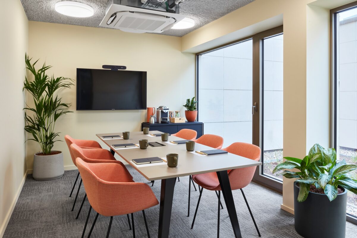 Een moderne conferentieruimte met een witte tafel omringd door zes oranje stoelen, een aan de muur gemonteerde tv en een console met een koffiezetapparaat. Grote ramen laten natuurlijk licht binnen en potplanten sieren de kamer.