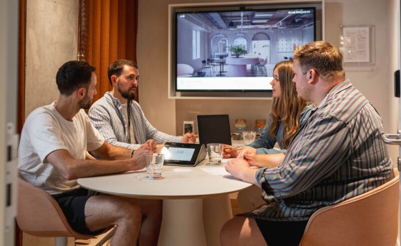 Four people sit around a small round table in a meeting room, discussing. A screen displays an interior design image behind them. They have documents, glasses of water, and a tablet on the table.
