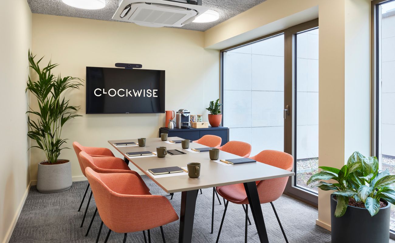 A modern conference room with a rectangular table surrounded by six orange chairs. A TV on the wall displays