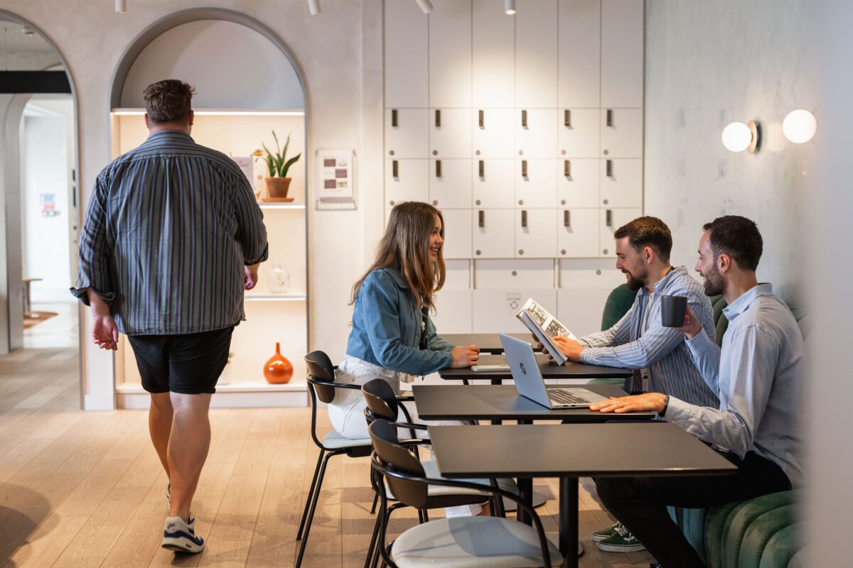 Drie mensen zitten met hun laptop en koffie aan een tafel, verwikkeld in een gesprek, in een moderne, lichte werkruimte. Een ander persoon loopt op de achtergrond weg naar zijn witte kluisjes.