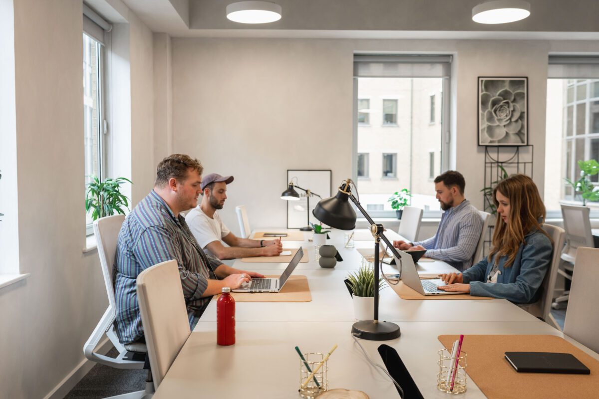 Four people work on laptops at a modern office table. The room has large windows, plants, desk lamps, and minimalistic decor. Two men and one woman sit on the right, another man sits on the left.