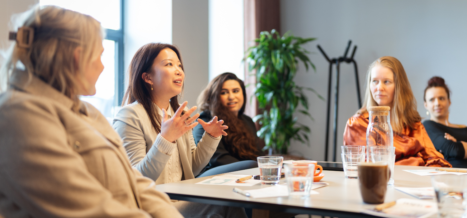 A group of women sit around a table in discussion, with one woman speaking animatedly. Papers, glasses of water, and a coffee are on the table in a bright, modern meeting room.