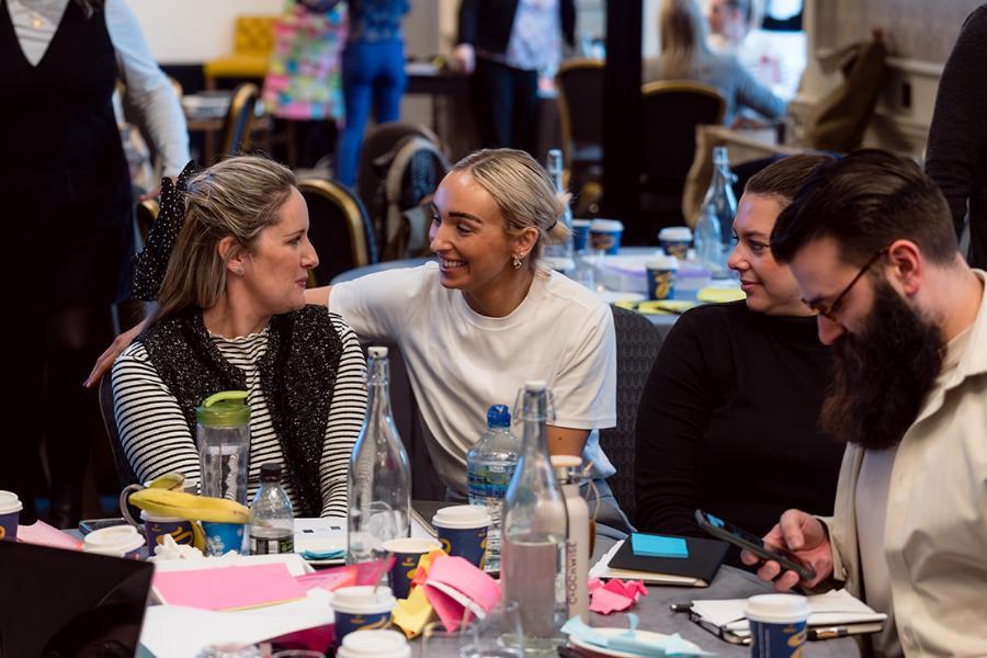 Four people sit around a cluttered table with drinks, water bottles, and papers, engaged in conversation and smiling. One woman has her arm around another, while one man looks at his phone. The atmosphere appears collaborative.