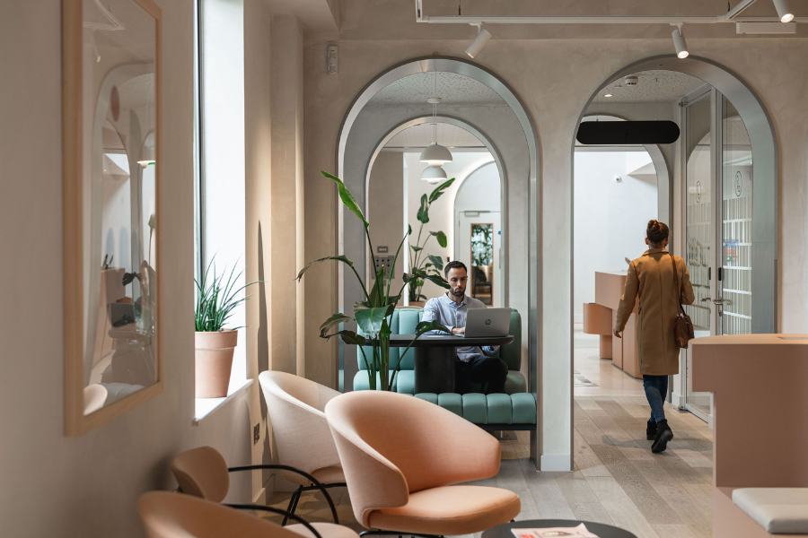 A man sits working on a laptop at a booth in a modern, stylish office with arched doorways, indoor plants, pastel chairs, and soft lighting. A person in a tan coat walks away in the background.