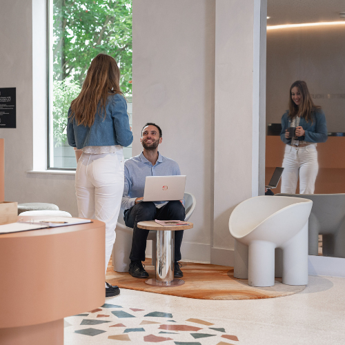 A man sits with a laptop on his lap, smiling up at a standing woman in a modern, light-filled office. The woman is reflected in a large wall mirror behind them. Contemporary furniture completes the setting.