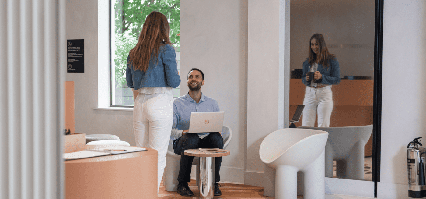 A man sits with a laptop on his lap, smiling up at a standing woman in a modern, light-filled office. The woman is reflected in a large wall mirror behind them. Contemporary furniture completes the setting.
