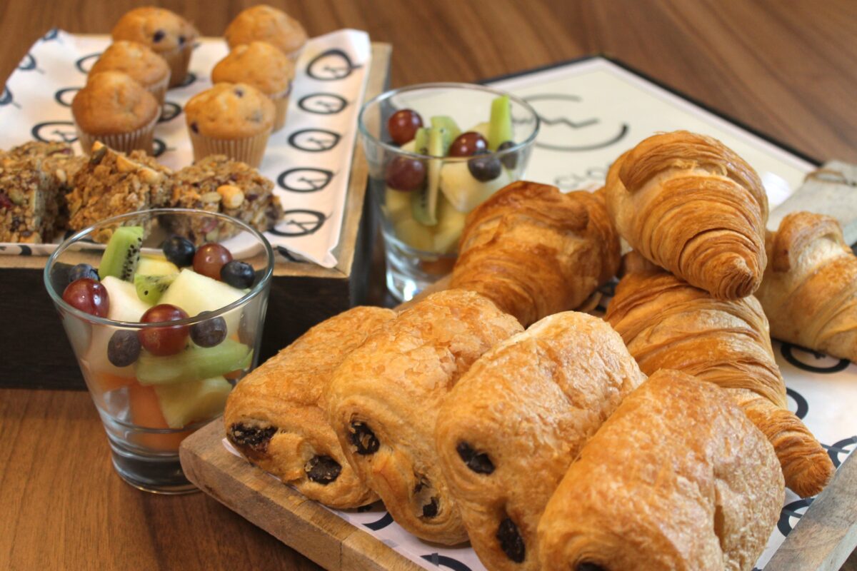 A wooden table with assorted pastries, including croissants and pain au chocolat, two glasses of mixed fruit, granola bars, and mini muffins displayed on trays and boards.
