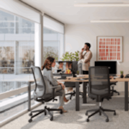 A modern office with large windows, two people present; one woman sitting at a desk using a computer, and one man standing in the background talking on the phone. The space is bright with contemporary furniture.