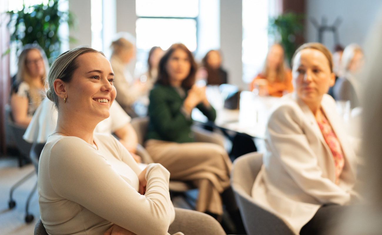 A group of women sit in a bright, modern room, attentively listening and smiling during a meeting or workshop. The focus is on a woman in the foreground smiling with her arms crossed.