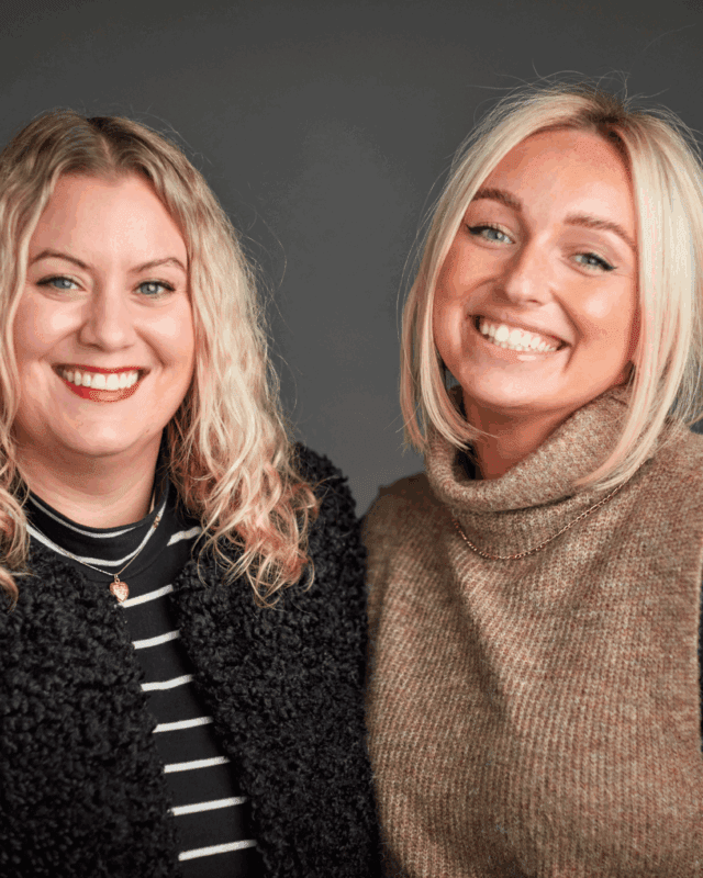 Two women with blonde hair smiling at the camera; one wears a black textured jacket over a striped top, and the other wears a brown turtleneck sweater. The background is a plain, dark gray.