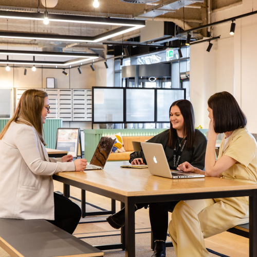 Three women sit at a wooden table in a modern, open office space, each using a laptop. They appear to be having a discussion or meeting. The room is bright with large windows and contemporary decor.