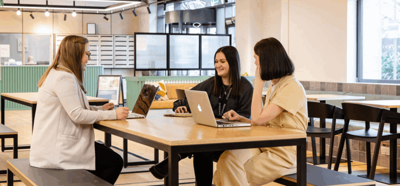 Three women sit at a wooden table in a modern, open office space, each using a laptop. They appear to be having a discussion or meeting. The room is bright with large windows and contemporary decor.