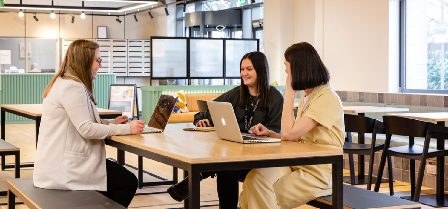 Three women sit at a wooden table in a modern, open office space, each using a laptop. They appear to be having a discussion or meeting. The room is bright with large windows and contemporary decor.