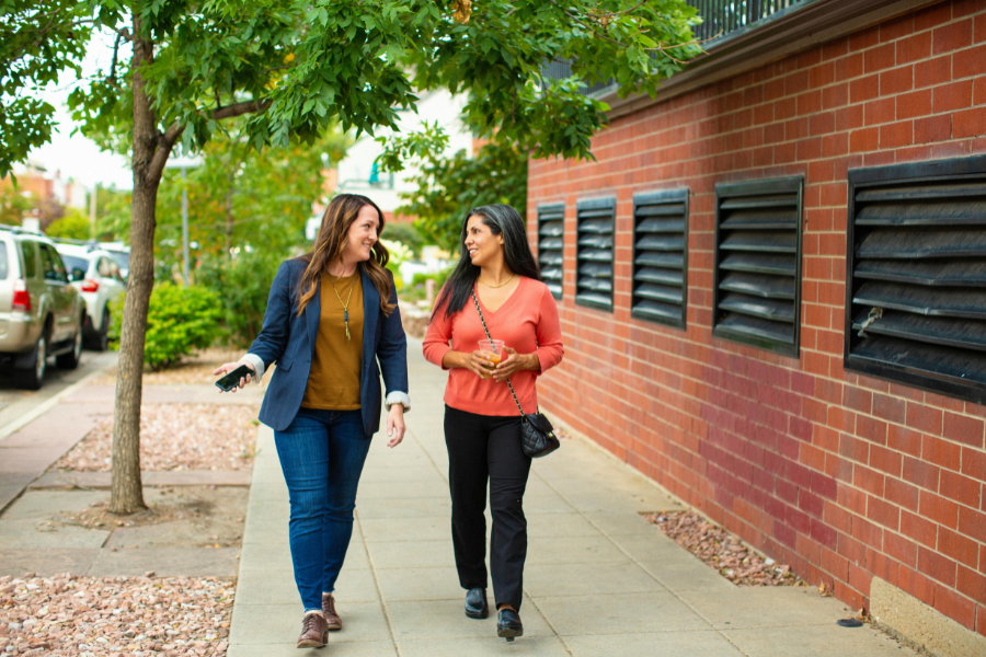 Two women walk and talk on a city sidewalk beside a brick building and trees. Both are smiling; one wears a blazer and jeans, the other a coral sweater and carries a black purse. Cars are parked along the street.