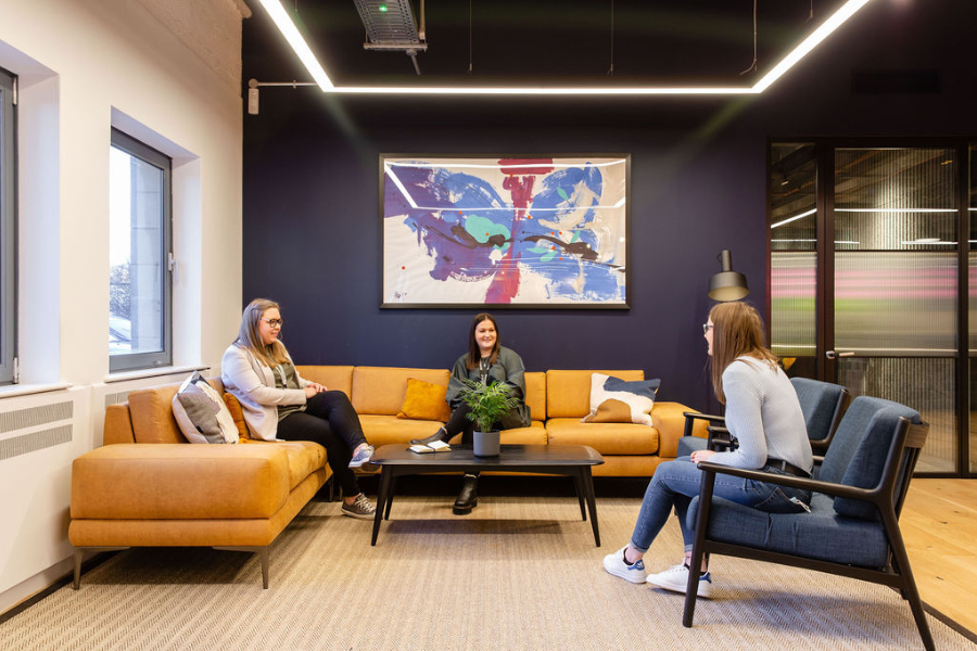 Three women sit and talk in a modern, brightly lit office lounge with a yellow sofa, blue chairs, a coffee table, and abstract artwork on a dark wall. Large windows provide natural light.