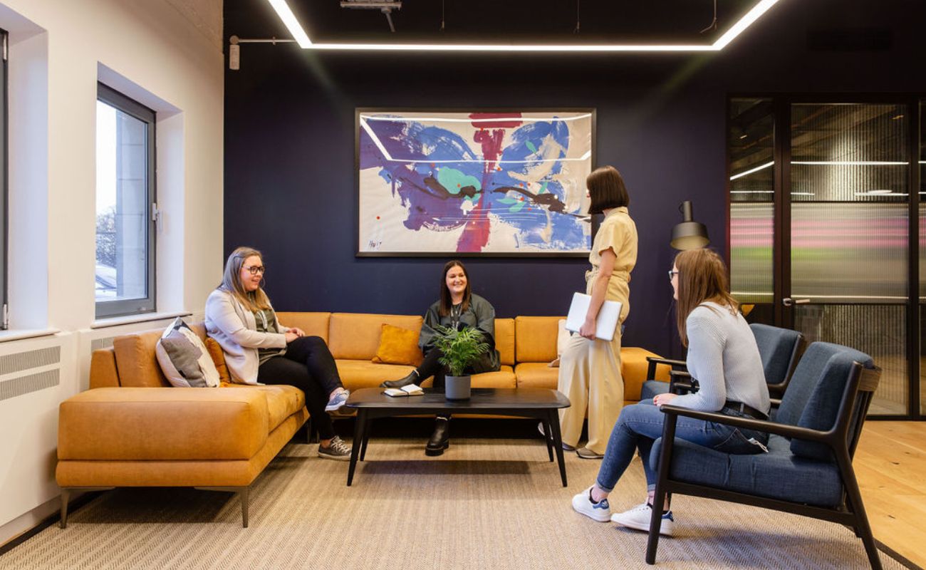 Four women are having a conversation in a modern office lounge with yellow sofas, a coffee table, and a large butterfly artwork on the dark wall. One woman stands holding papers while the others are seated, smiling.