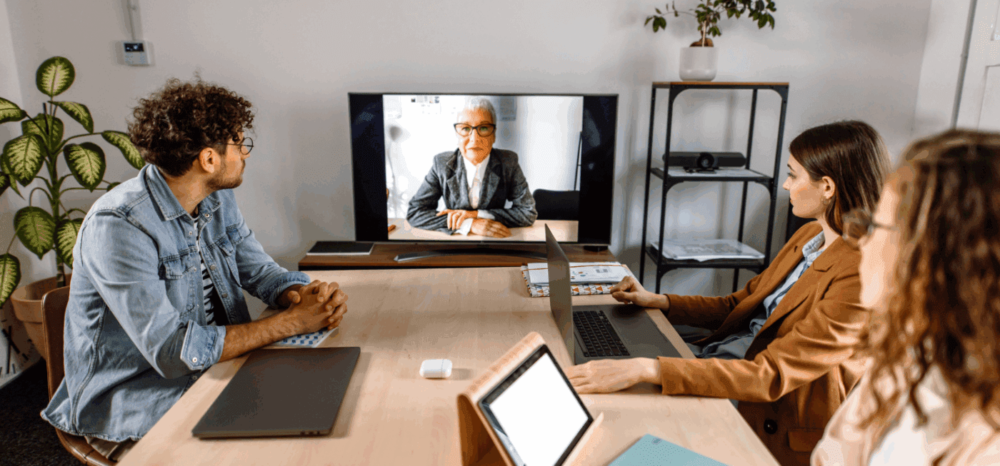 Three people sit at a conference table with laptops, attentively watching a woman speaking on a video call displayed on a large screen in a modern office setting.