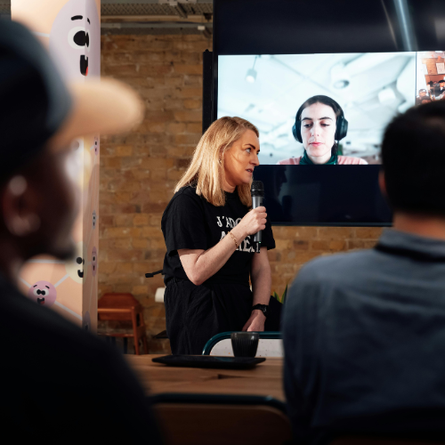 A woman speaks into a microphone in front of an audience, while another person wearing headphones appears on a large video screen behind her in a modern indoor setting.