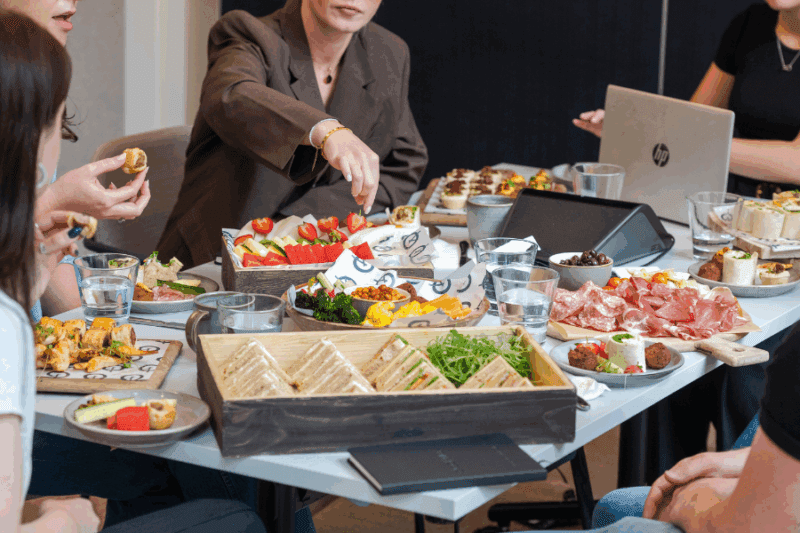 Four people sitting around a table share a variety of appetizers, sandwiches, fruit, and meats. Glasses of water, laptops, and plates are also visible, suggesting a casual business meeting or lunch gathering.