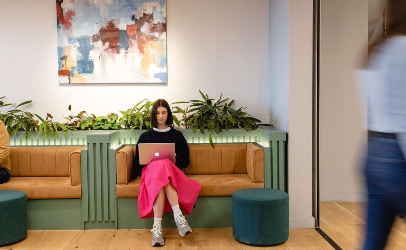A woman in a black top and bright pink skirt sits on a tan bench with green accents, using a laptop. Abstract art hangs above her, and plants line the wall. A blurred person walks past in the foreground.