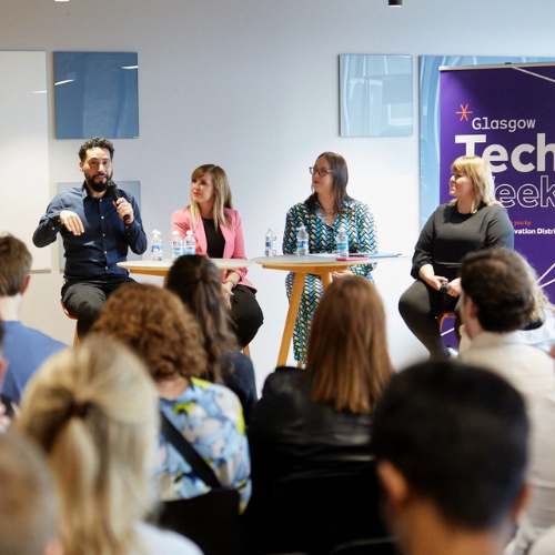A panel of four people speaks on stage at Glasgow Tech Week in front of an audience. One man is talking into a microphone, while three women sit beside him listening. A purple event banner is visible in the background.