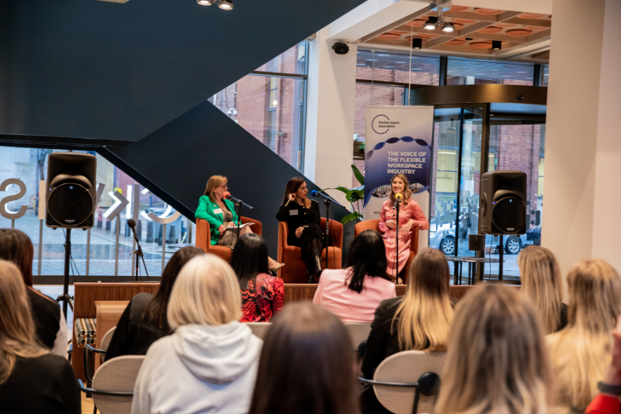 Three women sit on a stage speaking into microphones in front of an audience at an indoor event. Behind them is a banner that reads, 