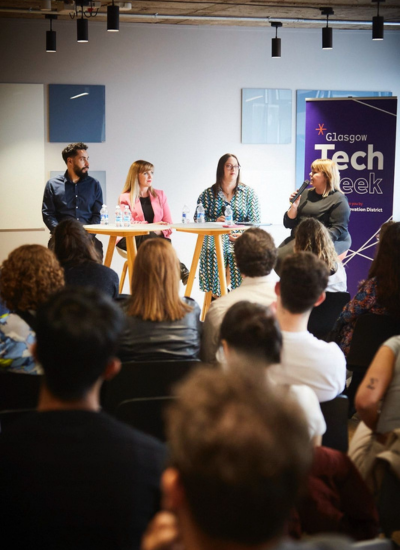 Four panelists sit at a table on stage, speaking to an audience at Glasgow Tech Week. One person holds a microphone, and a banner with the event name is visible in the background.