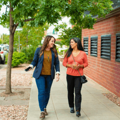 Two women walk and talk on a city sidewalk beside a brick building and trees. Both are smiling; one wears a blazer and jeans, the other a coral sweater and carries a black purse. Cars are parked along the street.