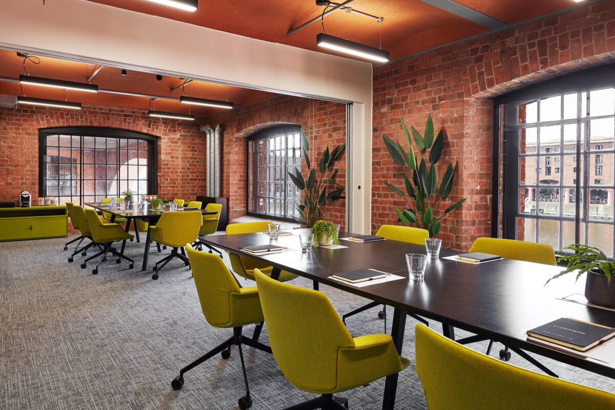 Modern meeting room with exposed brick walls, large windows, green chairs around black tables, potted plants, and notebooks with glasses of water on each table. Natural light fills the space.