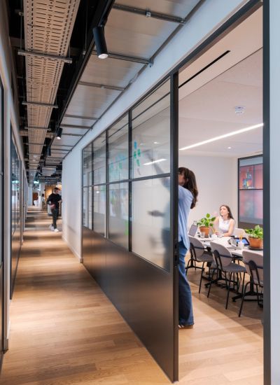 A modern office hallway with glass-walled meeting rooms; a person writes on a glass wall while others sit at a conference table with laptops and plants, and another person walks down the hallway.