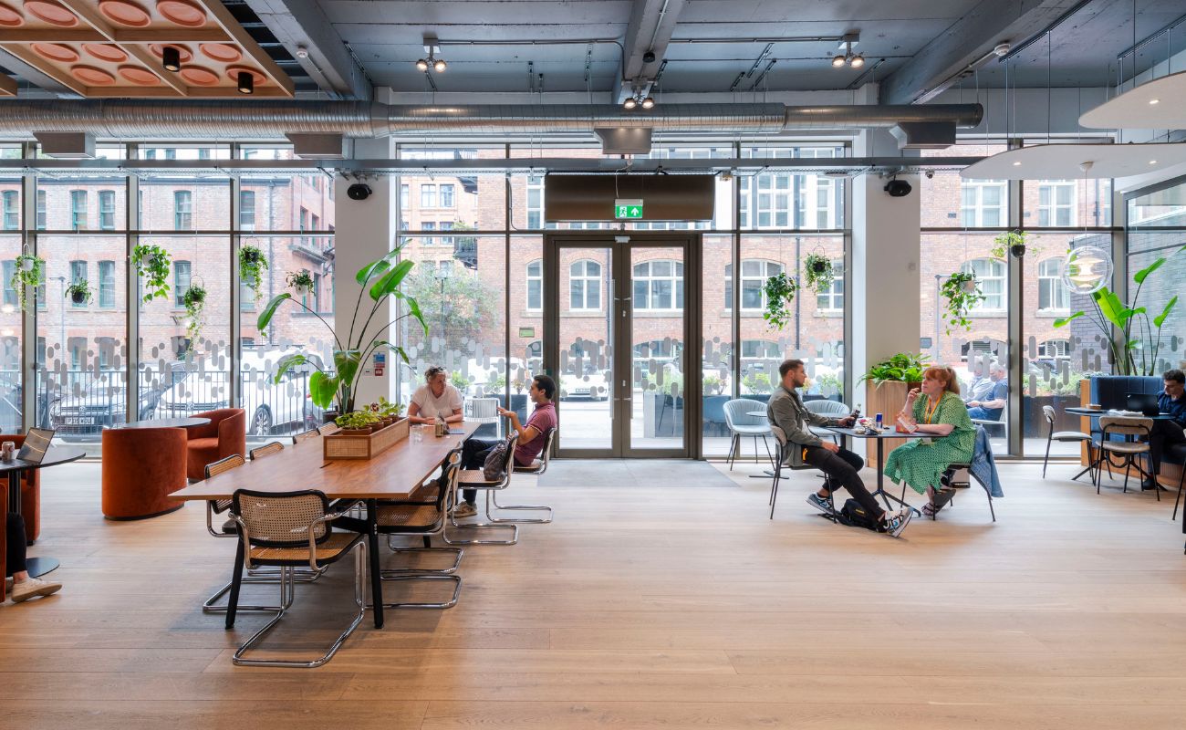 Modern open office space with large windows, indoor plants, wooden floors, and people sitting at tables talking or working on laptops. Red brick buildings are visible outside through the glass walls.