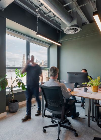 Two people sit at desks working on computers in a modern office with large windows and plants; a third person walks by, appearing blurred in motion. City buildings and a tower are visible outside the windows.