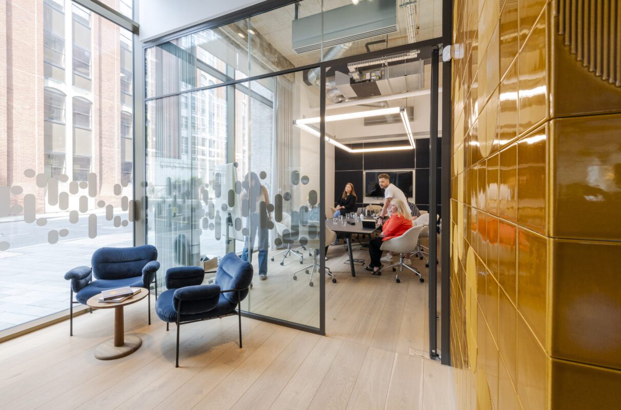Modern office with glass walls; three people sit at a conference table in a meeting room, while two blue chairs and a small table are in the lobby area. Yellow tiled wall is on the right. Large windows bring in natural light.