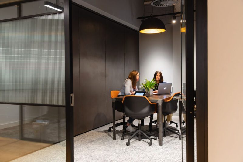 Two women sit at a table in a modern office meeting room, both using laptops and talking. The room has glass walls, a ceiling light, and a plant on the table.