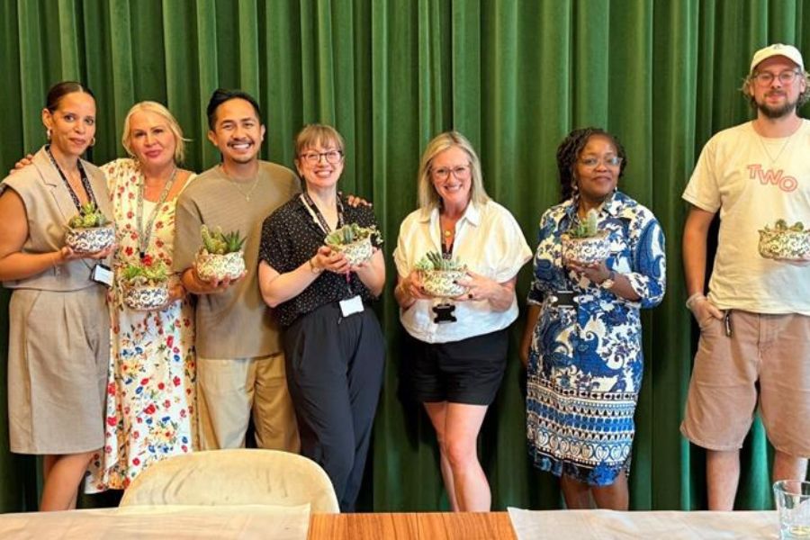 Seven people stand in a line indoors, smiling and holding small potted plants. A green curtain hangs in the background, and a table with light-colored tablecloths is visible in the foreground.