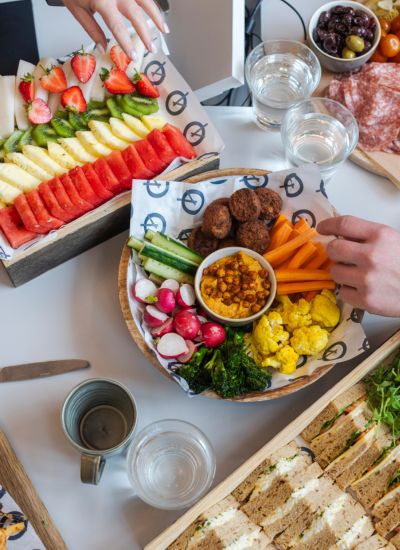A table set with plates of colorful fruit, fresh vegetables, falafel, pickles, olives, cured meats, and sandwiches. Two hands reach for food among glasses of water, mugs, and a variety of snacks.