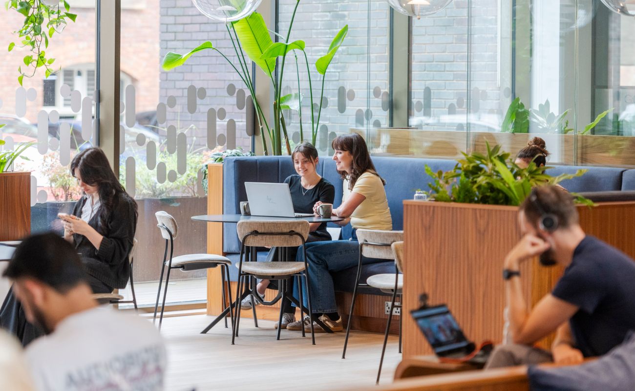 People work and socialize in a modern, bright cafe with large windows, lots of plants, and wooden furniture. Some use laptops, while others read or talk. The atmosphere appears relaxed and collaborative.