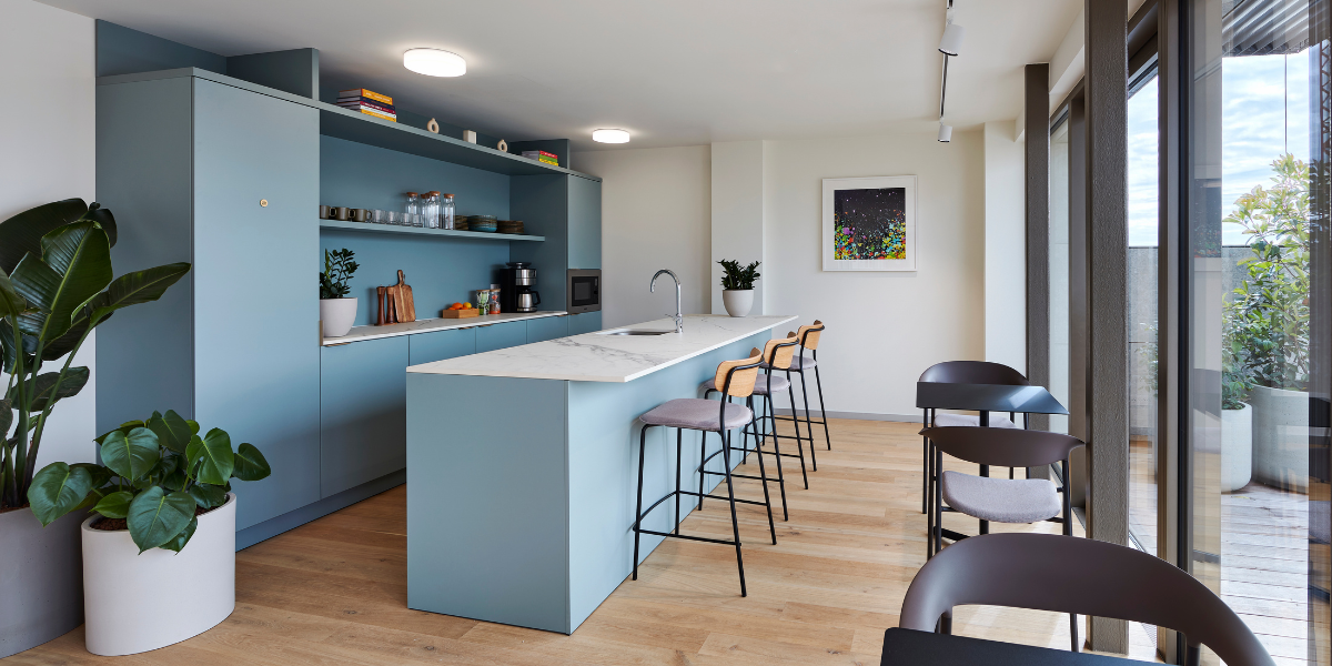 Modern kitchen with light blue cabinets, open shelves, a marble island with four bar stools, wooden floor, potted plants, wall art, and large windows letting in natural light.
