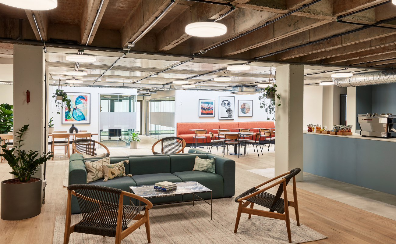 Modern office lounge with a green sofa, wooden chairs, and a coffee table on a rug. In the background, art hangs on white walls, with a dining area, plants, and a coffee counter visible. Exposed ceilings and large windows provide natural light.