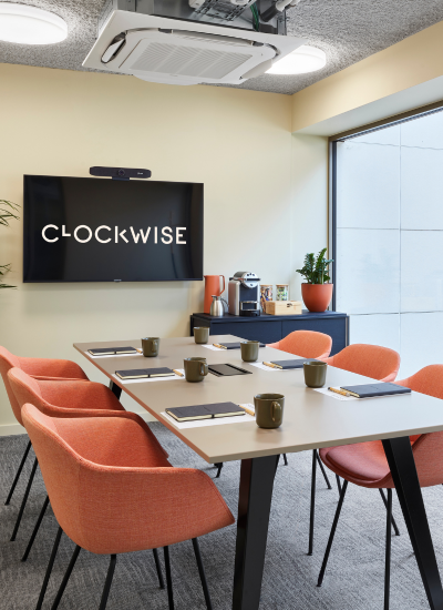 A modern meeting room with six orange chairs around a table set with notepads and mugs, a large wall-mounted screen displaying 