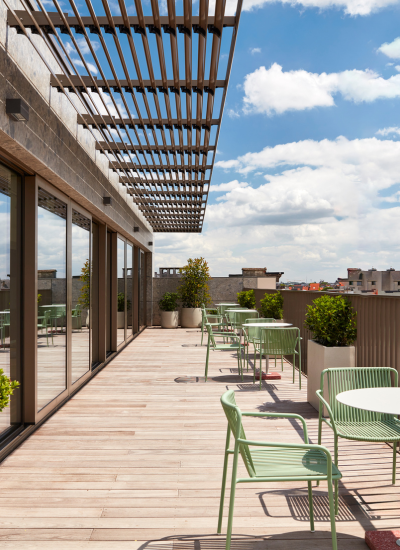 Rooftop patio with light green chairs and round tables, potted plants, wooden decking, and a pergola overhead. Large windows reflect the sky with scattered clouds and city rooftops in the background.