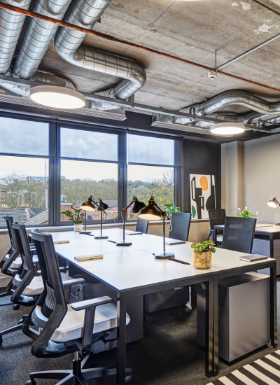 Modern office with large windows, several desks and ergonomic chairs, desk lamps, potted plants, and exposed ceiling ducts. Natural light fills the room, and abstract art hangs on the wall.