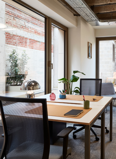 Modern office with two desks, ergonomic chairs, a tablet, stationery items, a lamp, and potted plants. Large windows let in natural light and offer a view of a brick wall and outdoor greenery.