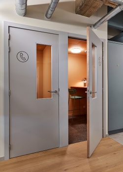 A modern office phone booth with a glass-paneled door slightly open, revealing a small workspace with a chair, desk, and overhead light inside. The door has a phone icon on it.